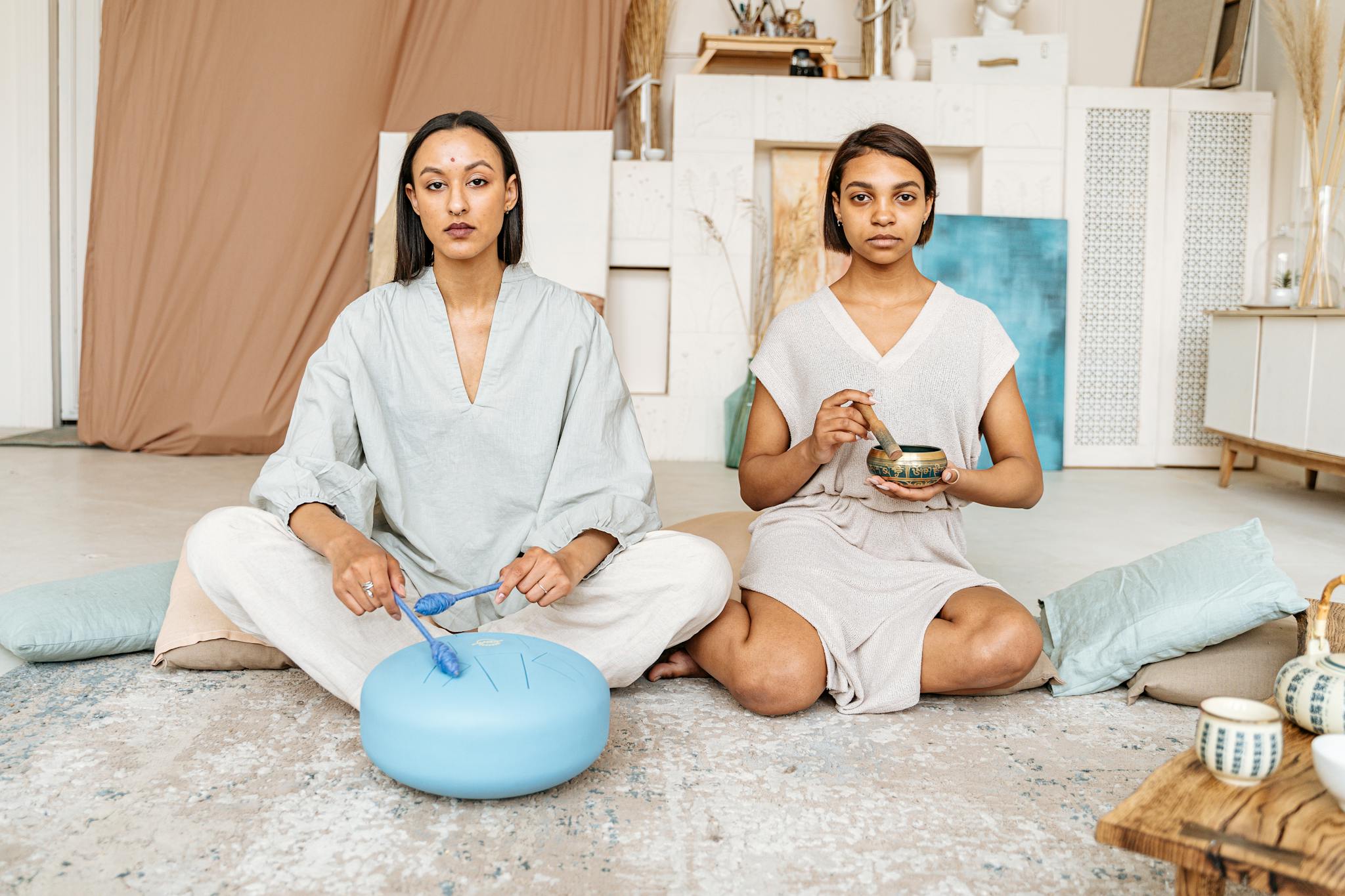 Two women meditating indoors with hang drum and Tibetan singing bowl, focusing on mindfulness and wellness.